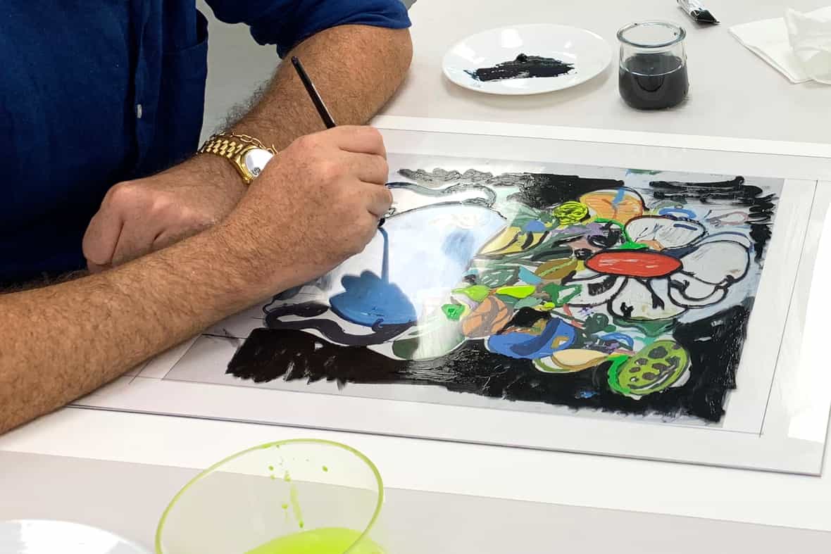 Artist Eddie Martinez sitting at a table while painting with a paintbrush on the surface of a clear plexi plate.