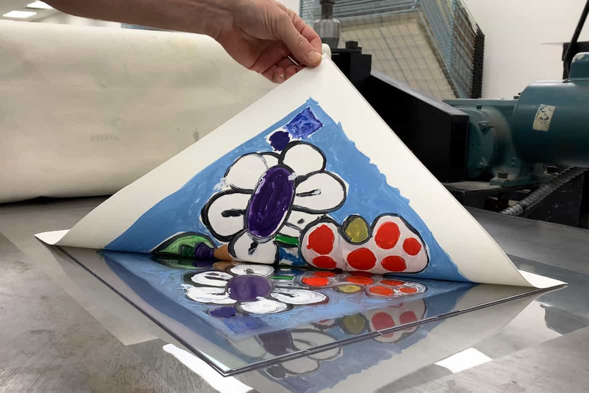 Close up image of a man pulling up the corner of a sheet of paper, revealing a printed image made using an etching press.