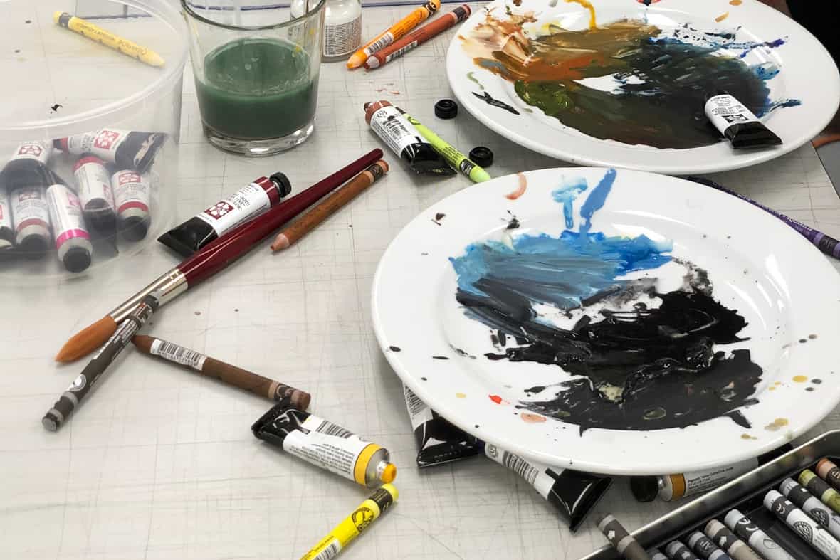 Close up image of a table with tubes of water color paints, brushes, and plates with paint palettes.