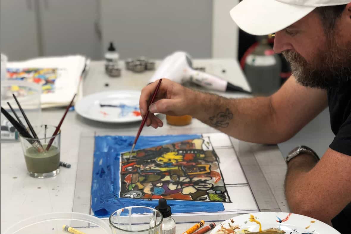 Artist Eddie Martinez wearing a white baseball cap and painting with a paintbrush on the surface of a clear plexi plate.